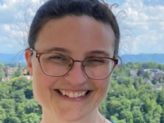 Kay wears glasses and a teal shirt, smiling in front of a green landscape with a blue sky and fluffy clouds