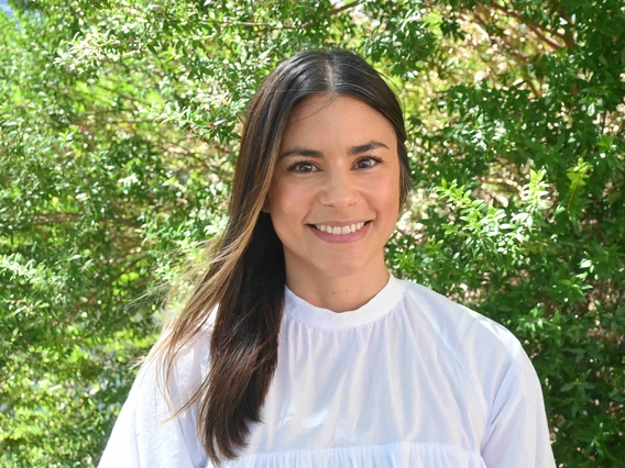 Melinda smiles in a white long-sleeve blouse in front of green trees on a sunny day.