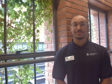 A smiling student in a black polo wearing a nametag stands before a brick wall and exterior window lined with dangling green vines